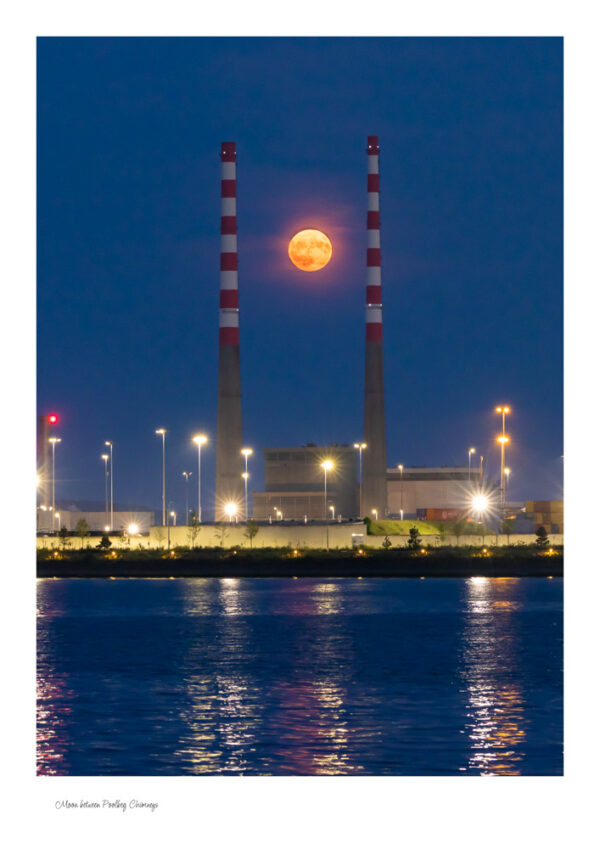 The Moon between Poolbeg Chimneys on a nice calm night with the reflections of the lights in the water